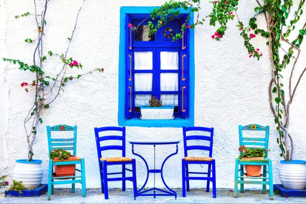 Vibrant blue wooden chairs and table in front of a quaint cafe with blue window frames and white walls, decorated with climbing roses.