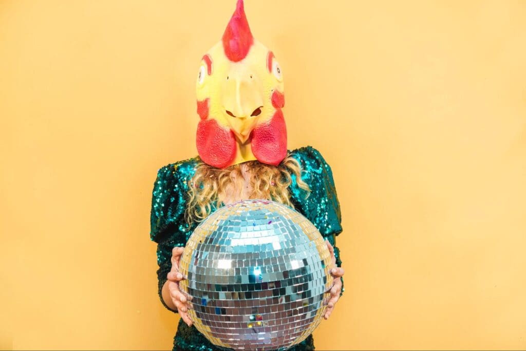Woman wearing a chicken mask holding a disco ball against a yellow background as one of the unique hen's party ideas.