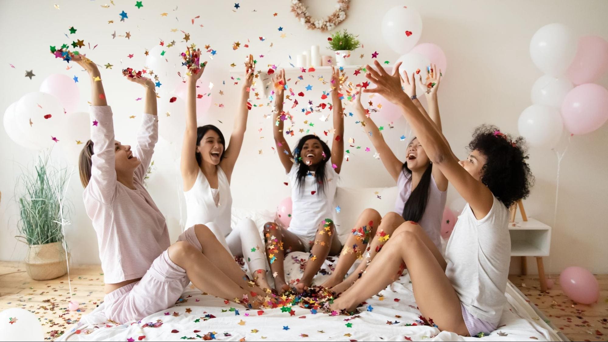 Group of joyful friends throwing confetti while sitting on a bed during a party, with balloons and festive atmosphere.