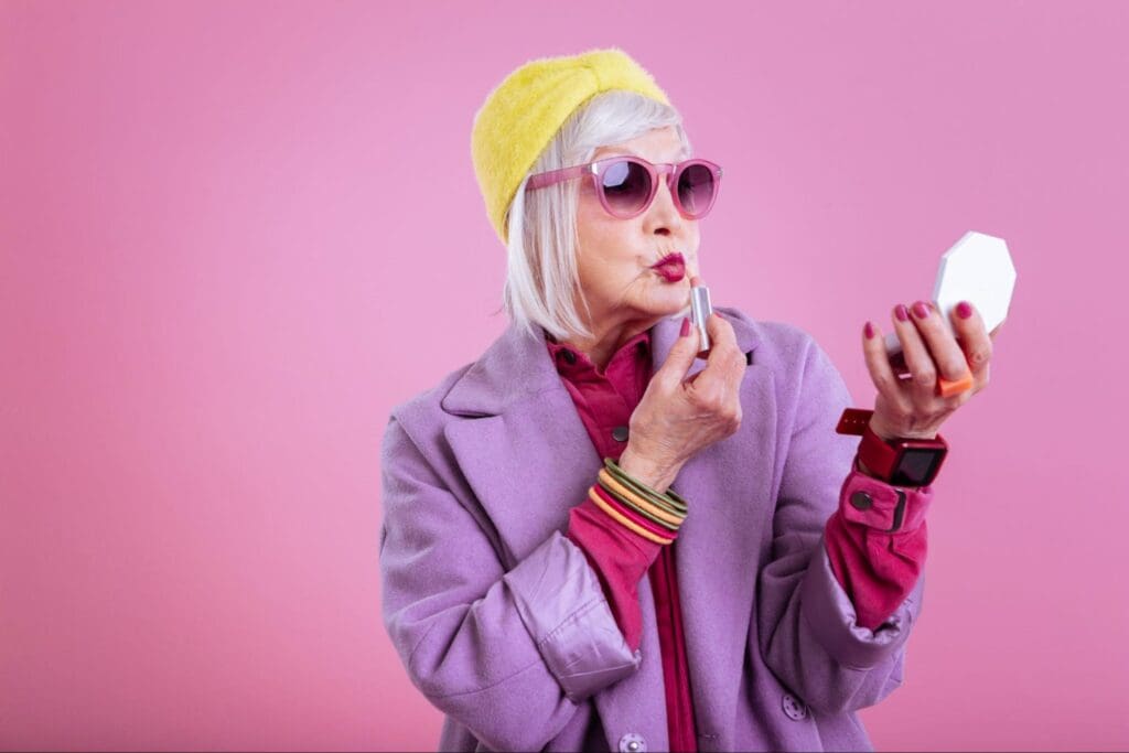 Elderly fashionable woman with a pink coat and yellow beret applying lipstick while looking into a handheld mirror.