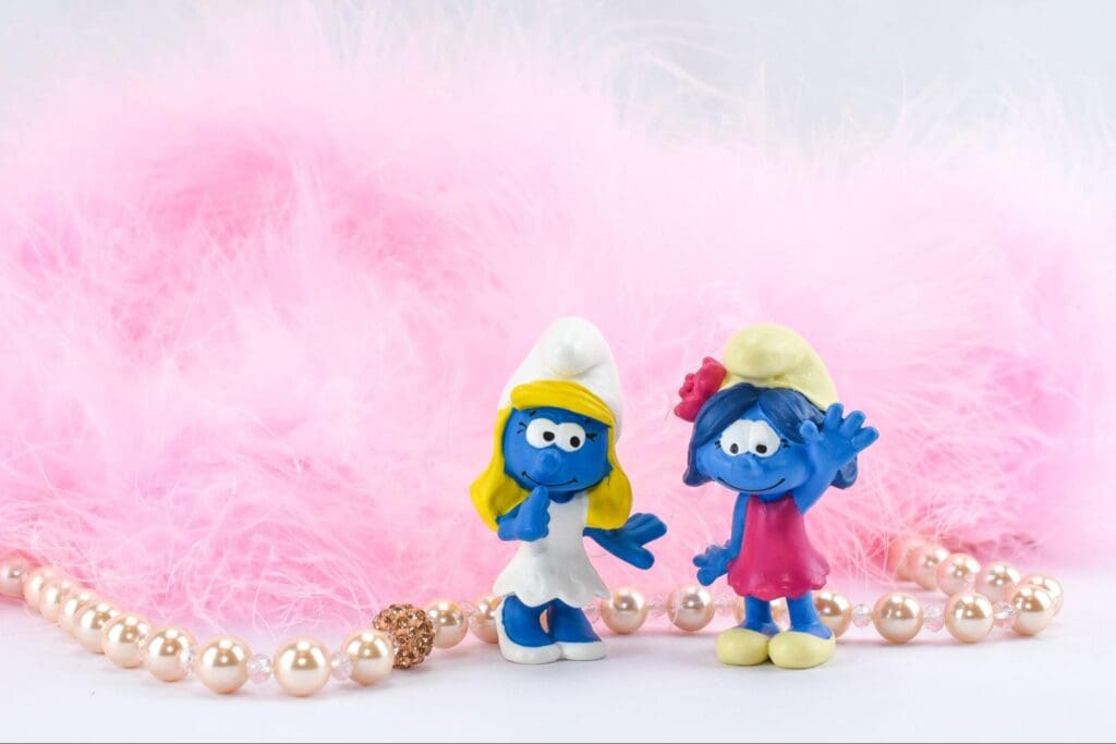 Two Smurf figurines, one blue and one pink, placed in front of a fluffy pink background with pearls.
