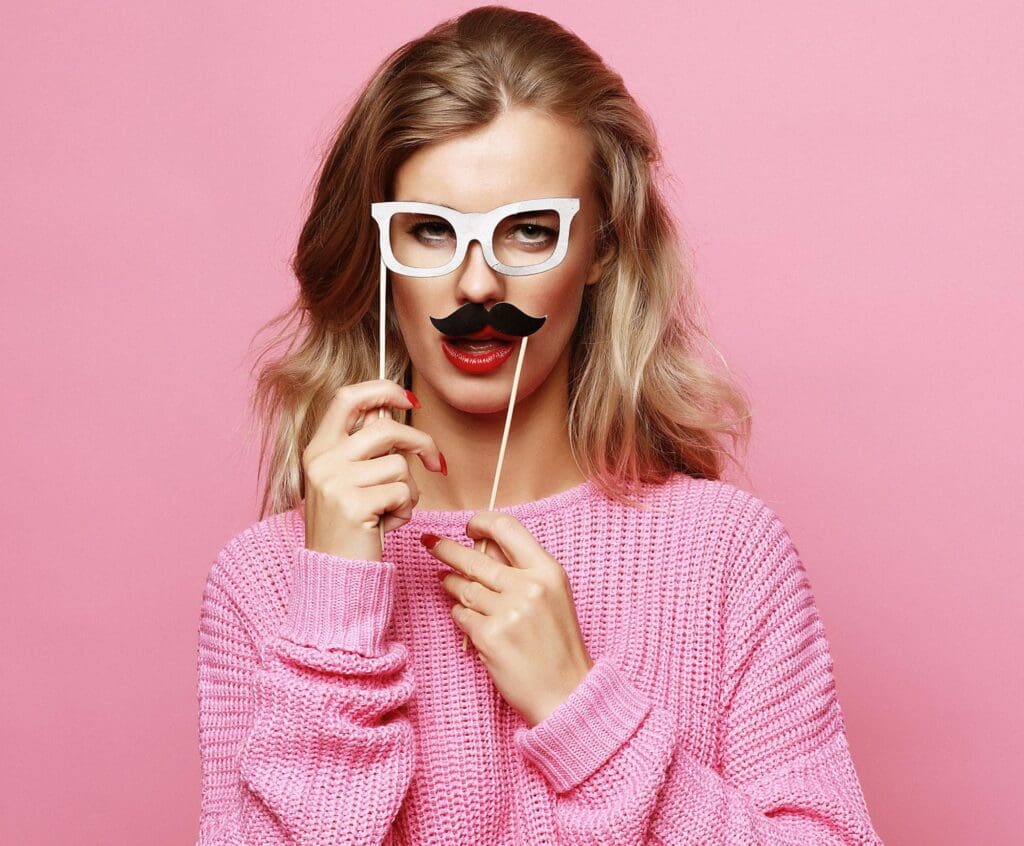 Young woman wearing a pink sweater and a white party mask shaped like glasses and mustache, holding the mask to her face and making a playful expression.