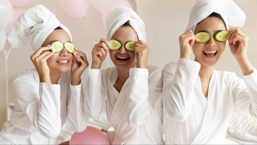 Three women wearing white robes and cucumber slices on their eyes, smiling and enjoying a pampering session at a pyjama party.