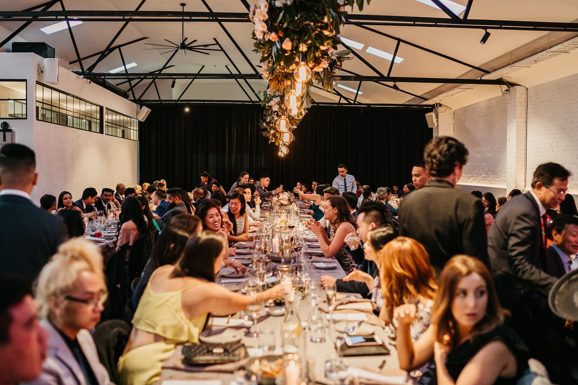 A large group of people seated at long tables, likely celebrating a wedding, in a spacious venue setting.