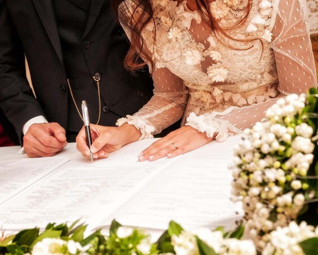 Bride in a lace dress signing a marriage certificate with the groom standing beside her, both focusing on the paperwork.