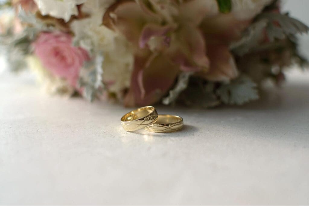 Two gold wedding rings placed on a table with a bouquet of flowers in the background, featuring pink and white roses.