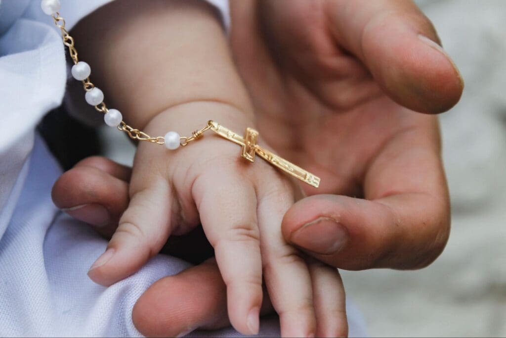 A close-up of an adult hand gently holding a baby's hand adorned with a delicate gold cross necklace and pearl bracelet.