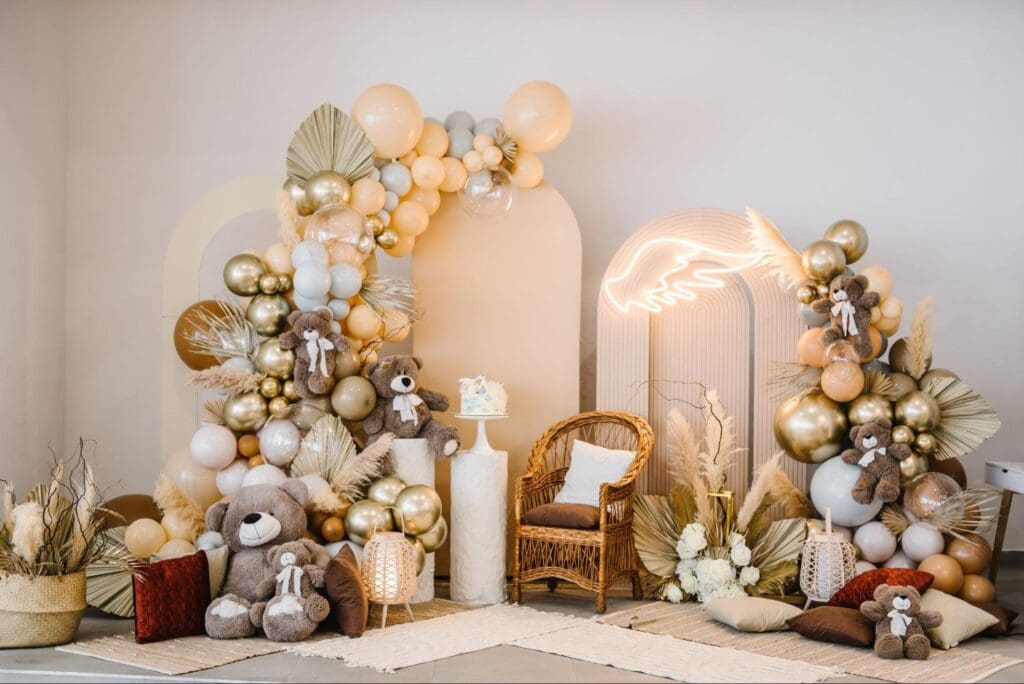 A beautifully decorated baptism party setup with pastel and gold balloons, teddy bears, a wicker chair, and a neon light shaped like a hand.