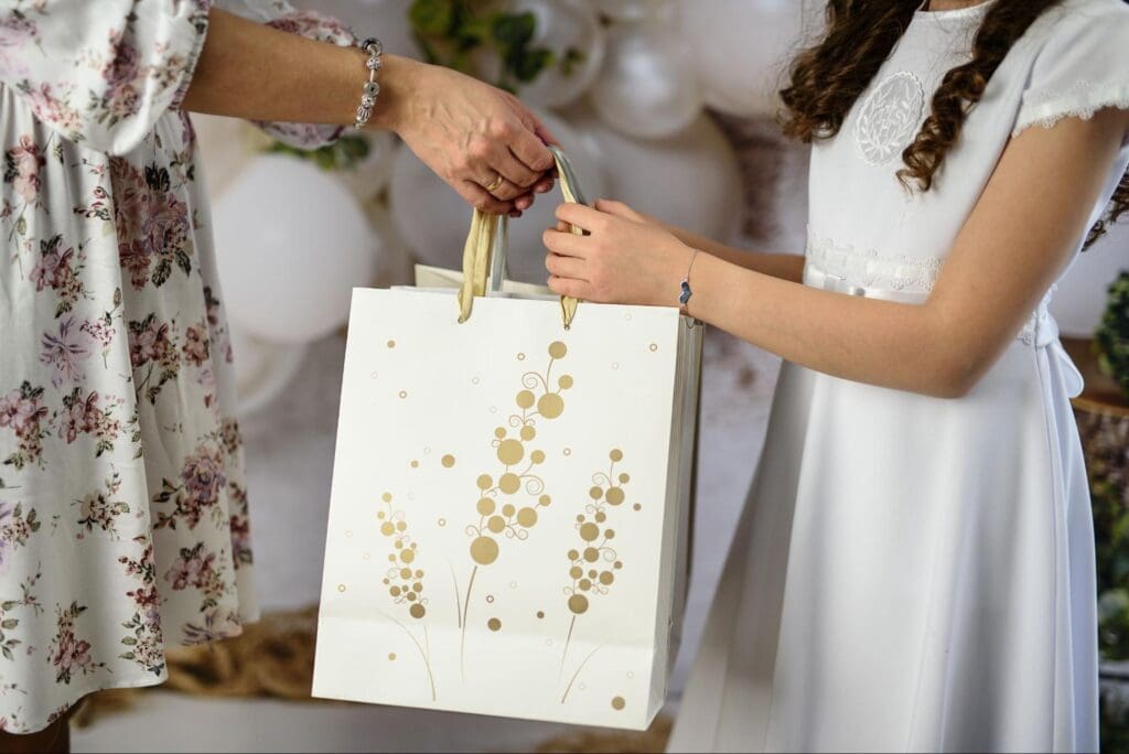 A young girl in a white dress receiving a gold and white gift bag from an adult wearing a floral dress during a special occasion.