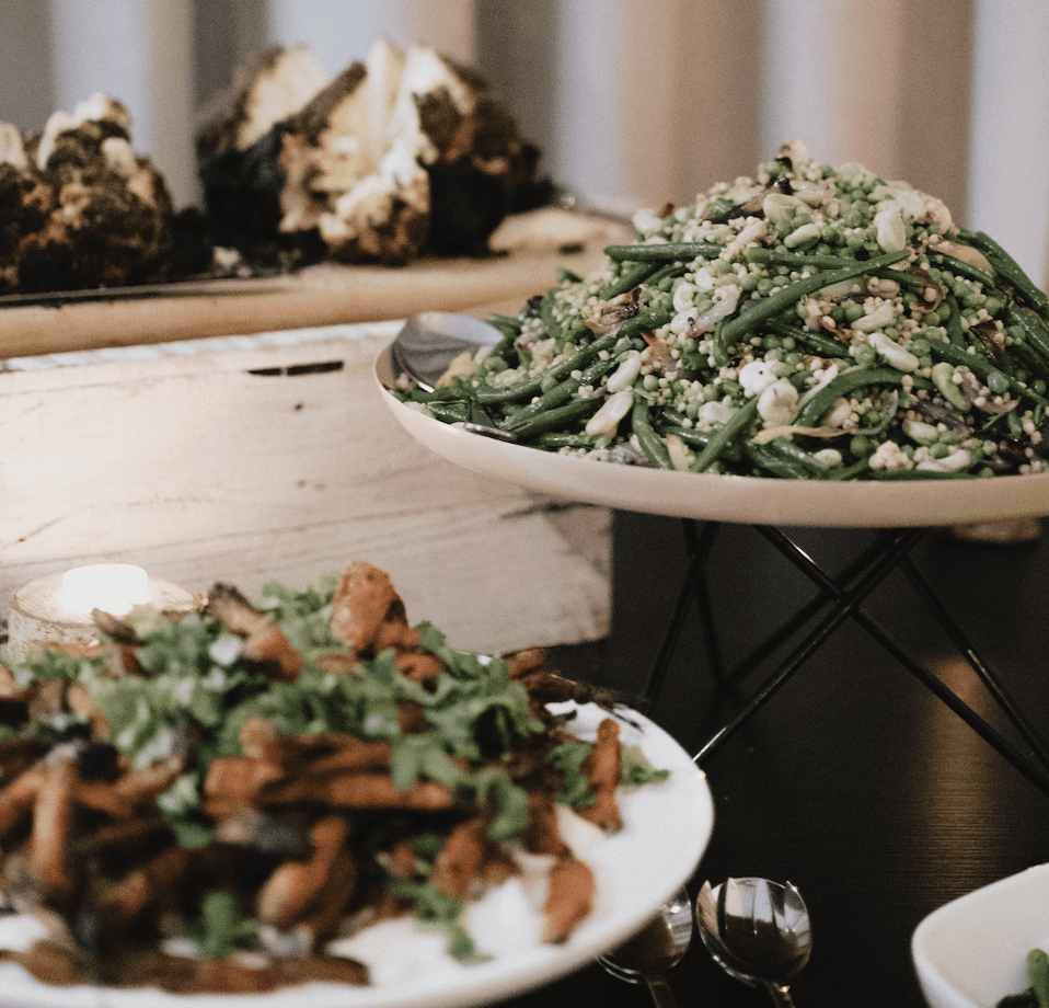 A buffet table featuring roasted carrots garnished with cilantro, green bean and couscous salad, and roasted cauliflower, set up with serving utensils.