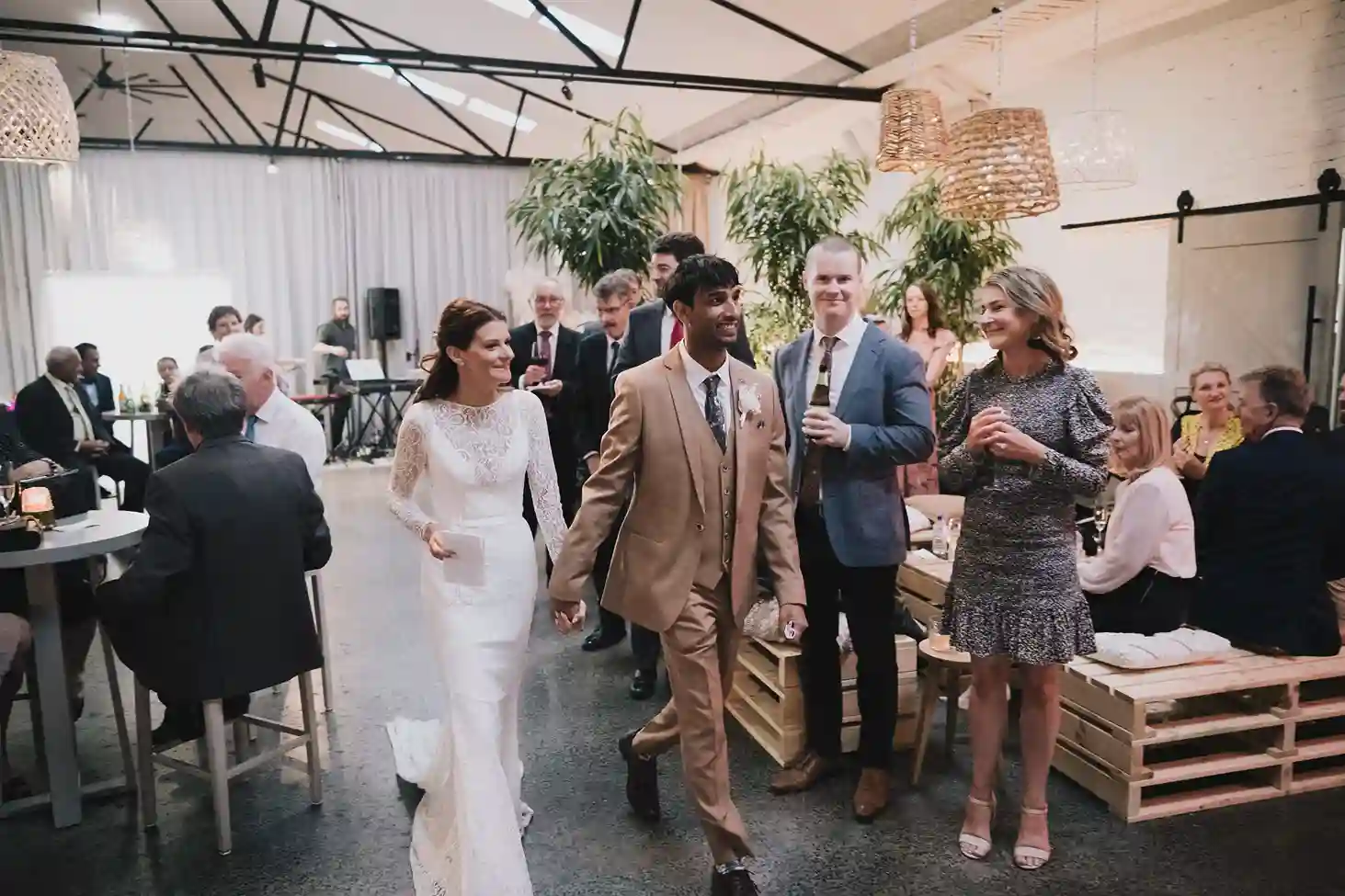 Bride and groom making their entrance at a wedding reception, surrounded by smiling guests in a warmly lit venue.