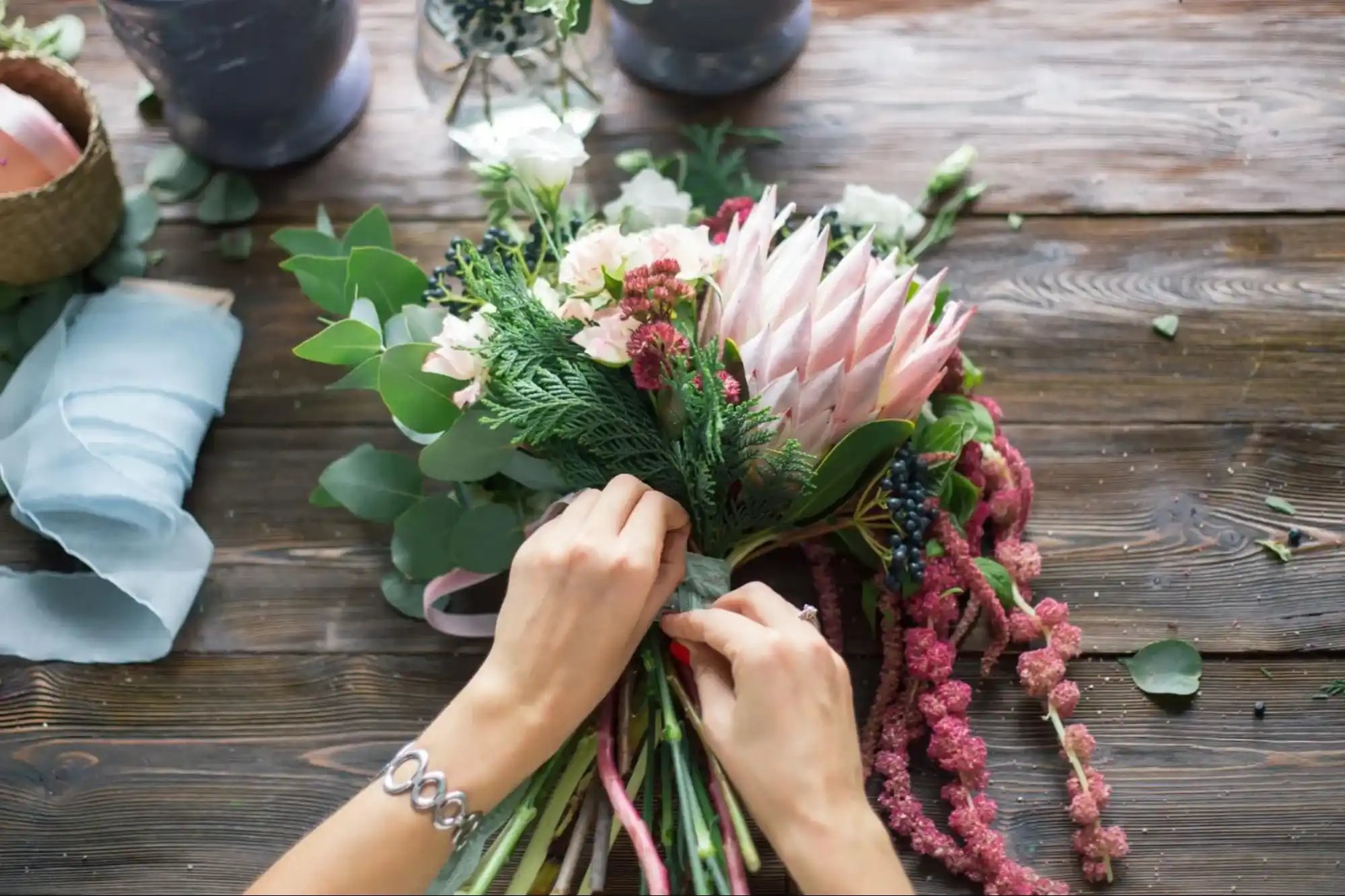 Hands arranging a bouquet of fresh flowers on a rustic wooden table, featuring vibrant pink proteas, greenery, and a soft blue ribbon.