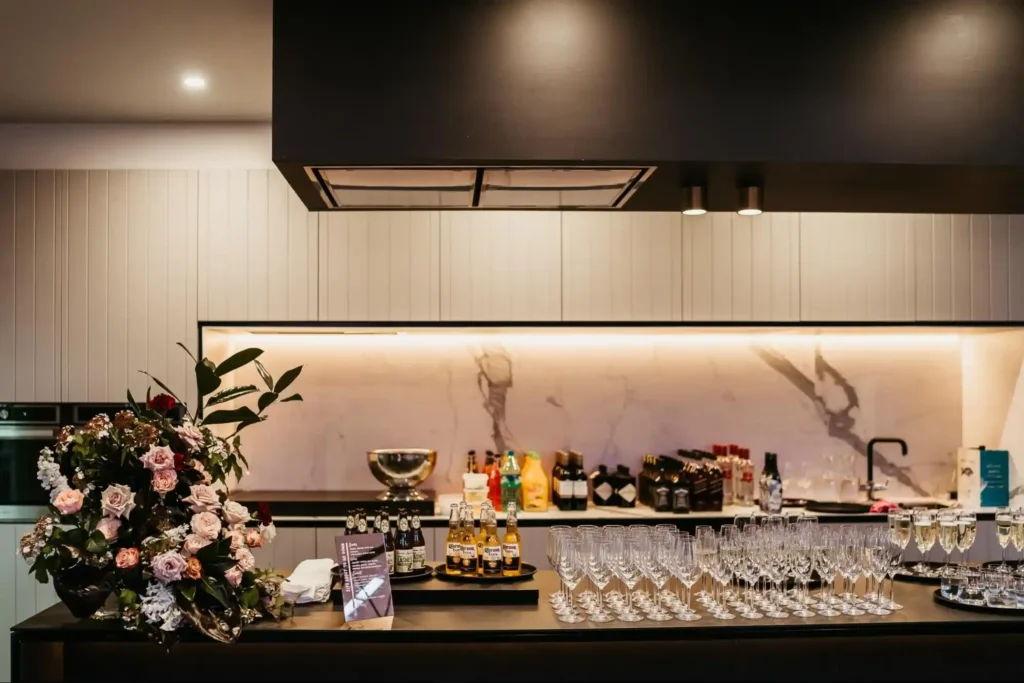 A luxury bar setup featuring an elegant floral arrangement, glassware ready for service, and bottles of beverages, set against a modern marble backsplash.