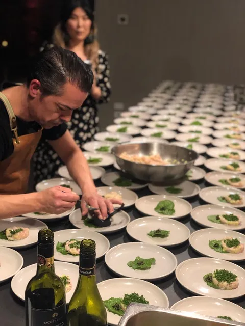 Chef carefully plating multiple dishes for a large dining event, with a row of prepared plates on the table.