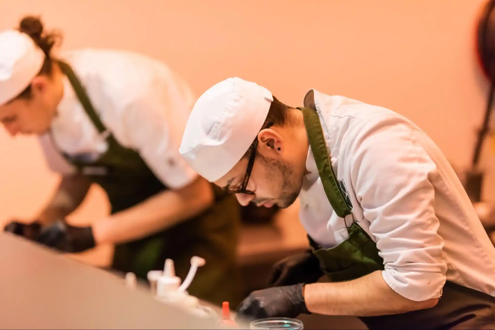 Two chefs wearing white hats and green aprons preparing food in a professional kitchen with focused expressions.