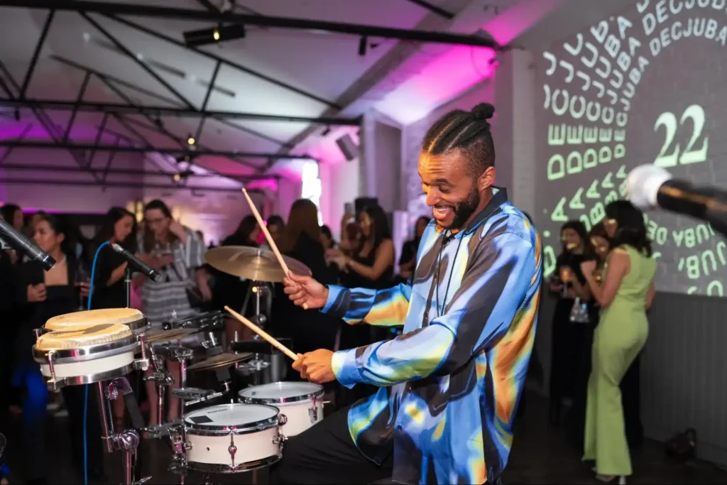 A smiling drummer performing live at a party, surrounded by guests in a vibrant event venue.