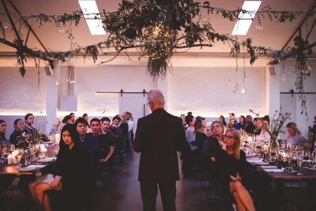 Man giving a speech to guests seated at an event dinner, with rustic ceiling decorations and a casual, elegant vibe.