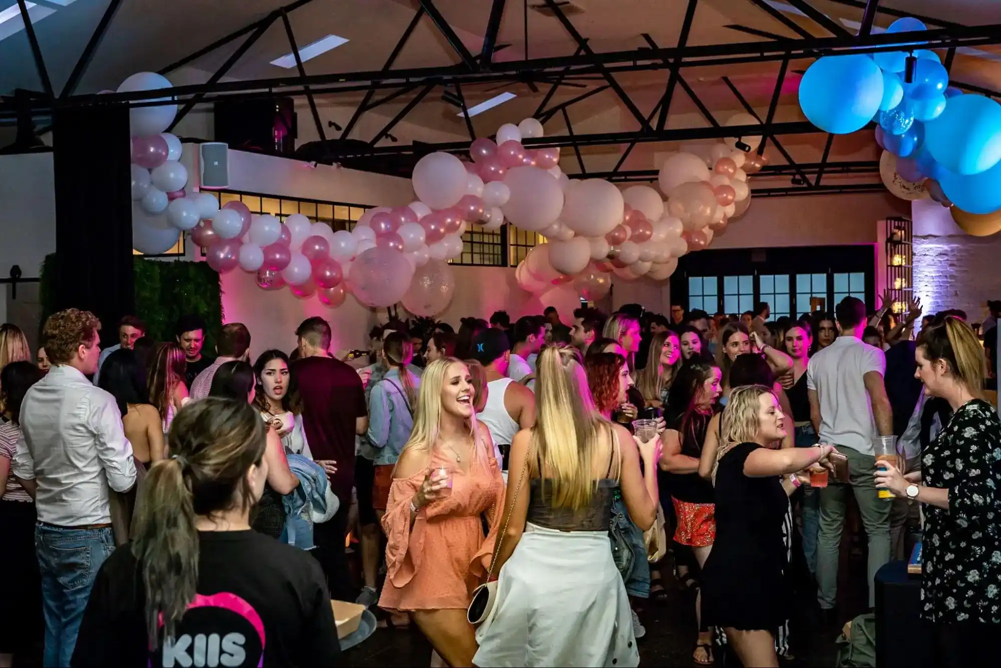 A lively party with guests mingling and dancing under pink and white balloon decorations in a stylish venue.