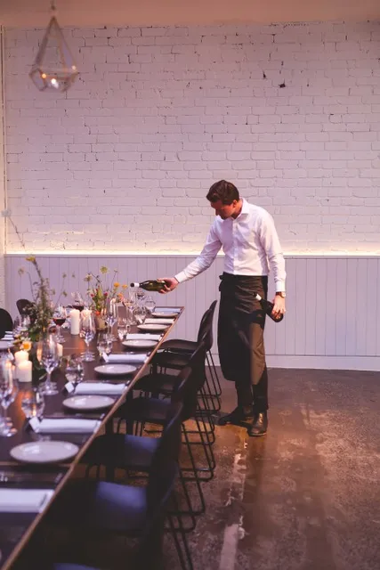 Waiter pouring wine into glasses at a long dining table, set in a minimalist venue with white brick walls.