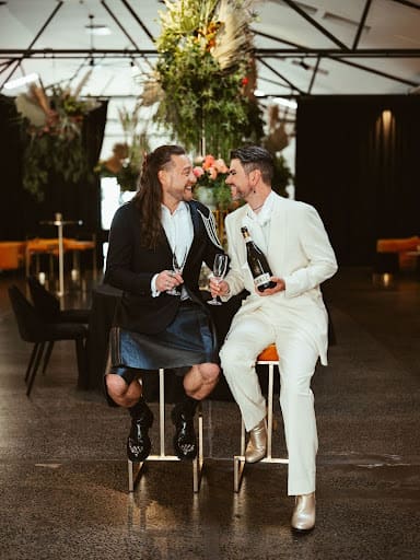 Aarron and Robert, dressed in suits, are seated on chairs, each holding a wine glass, appearing to share a toast.