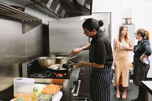 Chef preparing a dish in a kitchen while two women observe and engage in conversation nearby. 