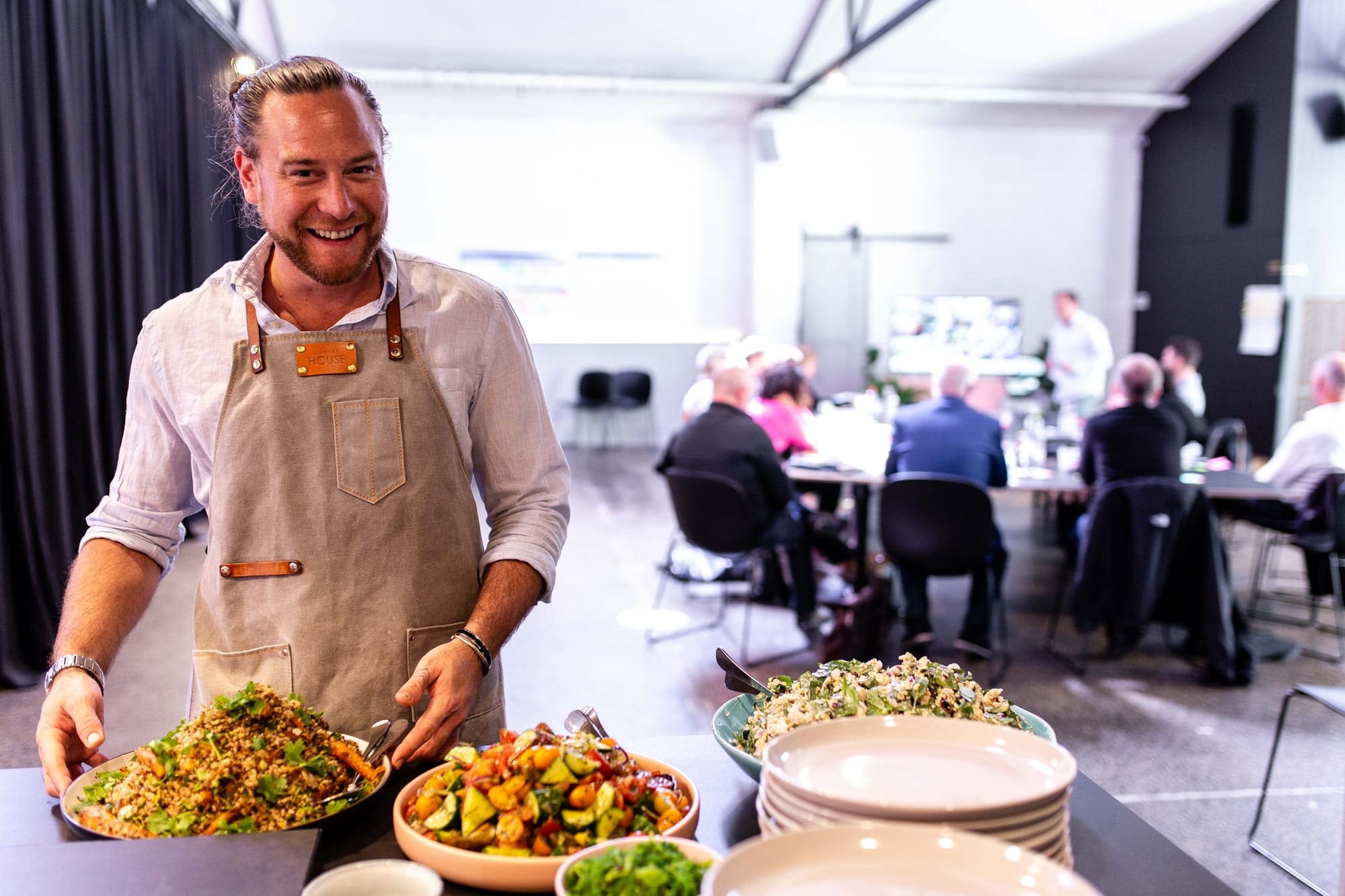 A man in an apron serves food at a table during a hybrid conference event in a physical venue.
